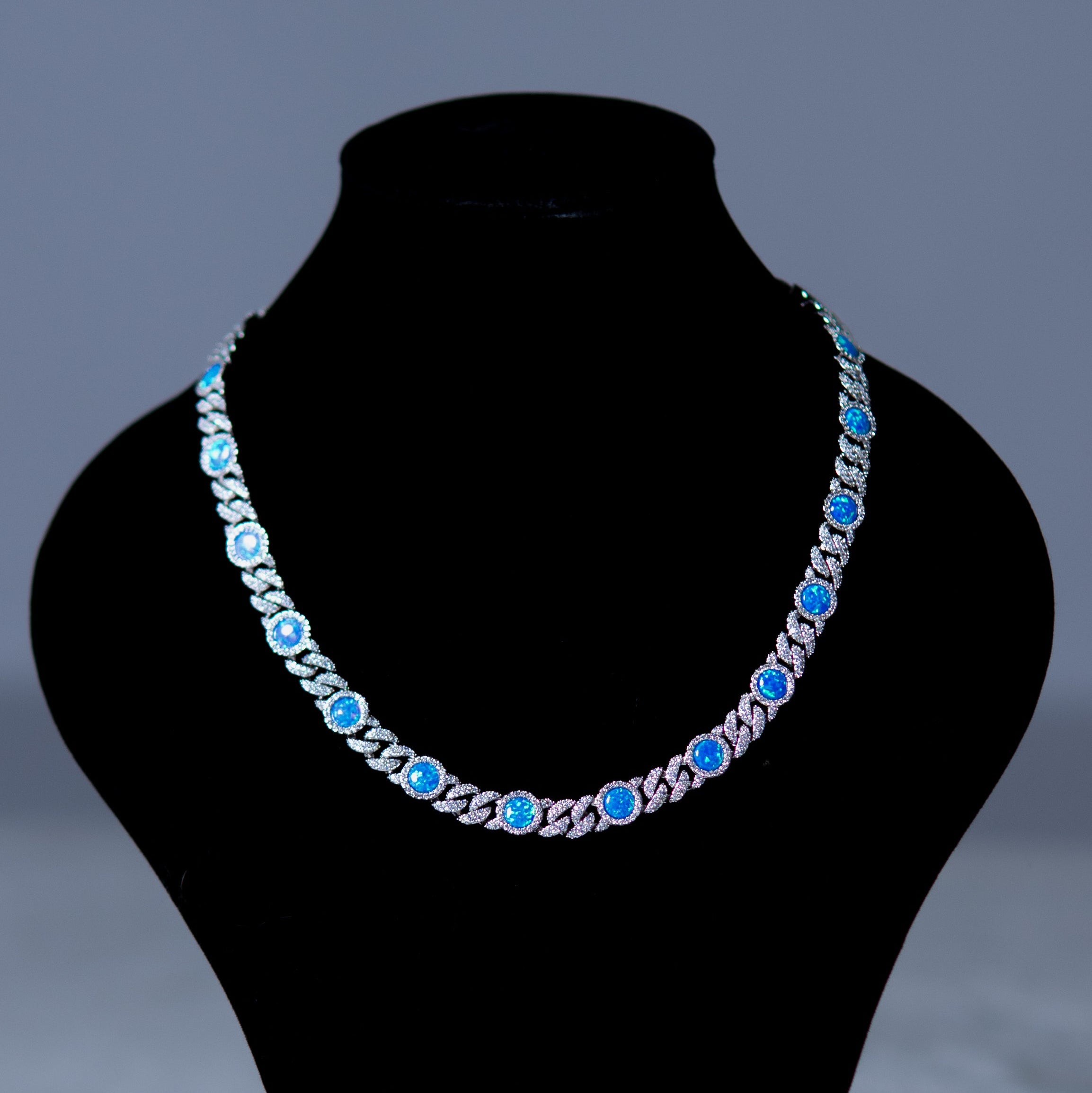A silver-toned chain necklace with round blue gemstones, displayed on a black jewelry bust against a dark gray background.