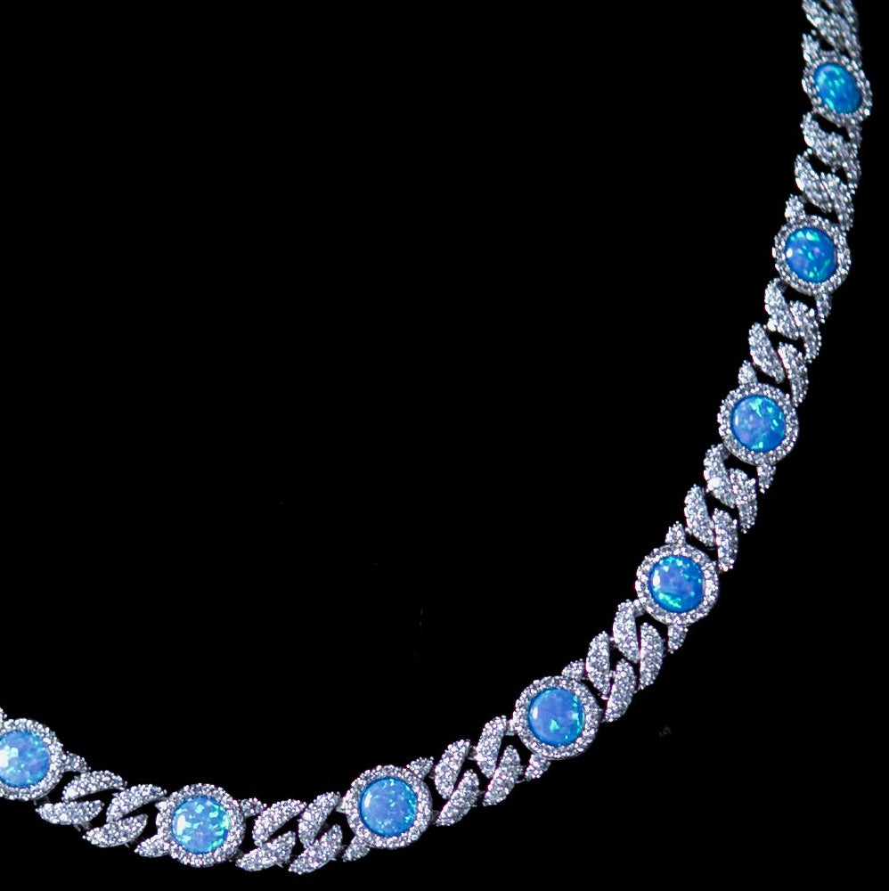 A silver-toned tennis necklace with round light blue gemstones and pavé accents, displayed on a black velvet jewelry bust against a dark gray background.