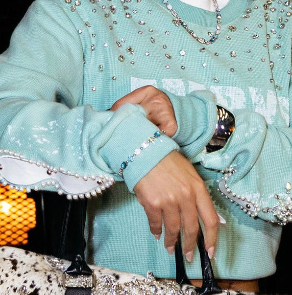 A woman wearing a light aqua blue embellished sweatshirt with white trim and rhinestone details, holding a black and white cow-print handbag in a dark indoor setting.